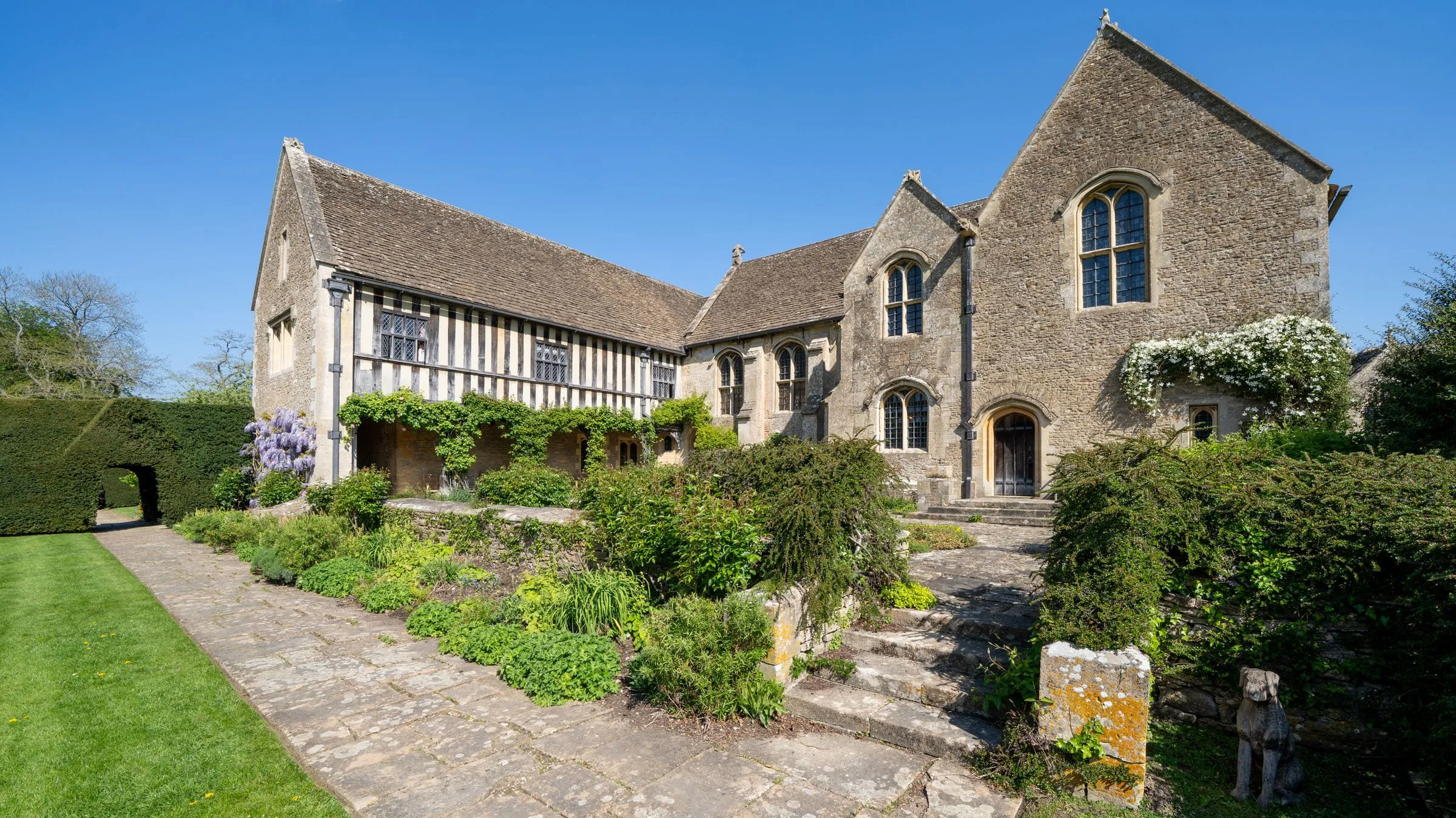 Wide exterior stock photograph of a historic English manor in mixed pale stone masonry, exact stone type unverified, shown at whole-building scale.