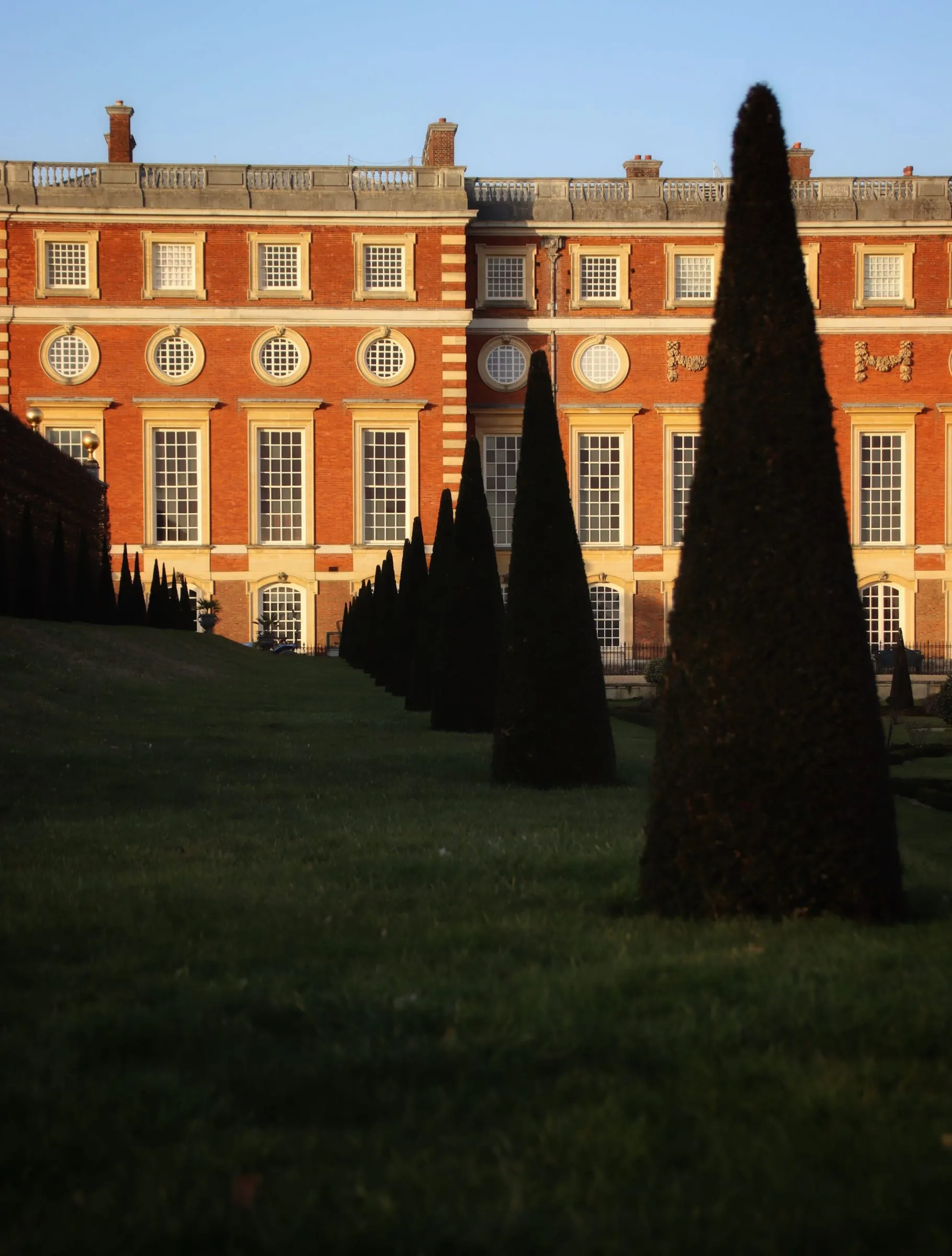 Portrait-format stock photograph of Hampton Court formal gardens and a red-brick facade with pale stone dressings, stone type unverified, shown at palace-and-landscape scale.