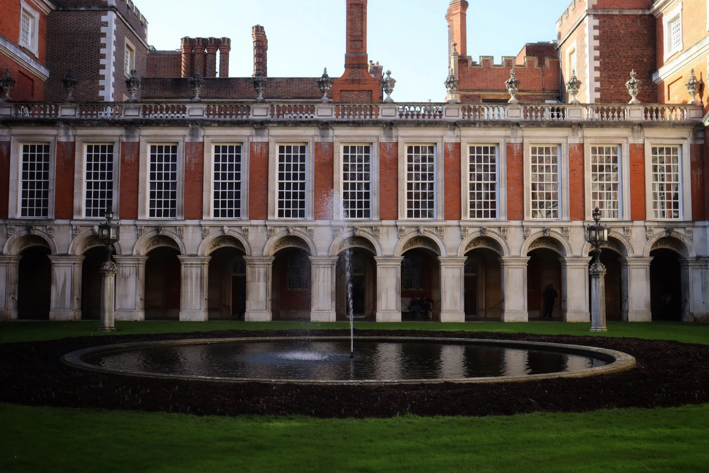 Wide stock photograph of a Hampton Court fountain and pale stone arcade, stone type unverified, shown at courtyard scale.